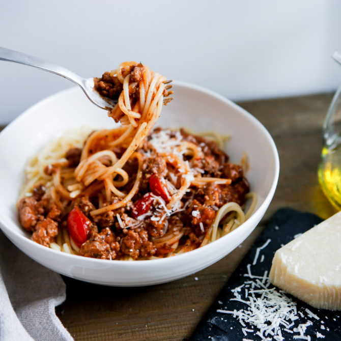 Valkoisessa kulhossa on spagettia ja jauhelihakastiketta, jota ihmisen käsi ottaa haarukalla. Kulhon vieressä on lasikannu jossa on öljyä ja musta liina, jonka päällä on pala parmesaani juustoa.