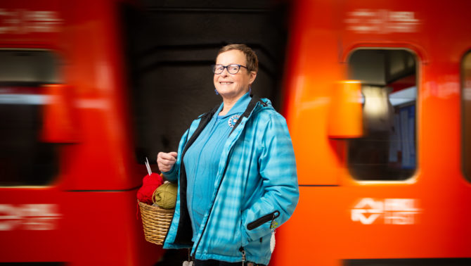 Nainen vaaleansinisessä takissa pitelee kädessään koria täynnä virkkaustarvikkeita. Hänen takana kulkee metro.