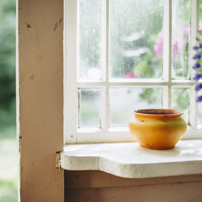 Valkoisella ikkunalaudalla on vaalean oranssi ruukku, jonka vieressä on violetteja kukkia. Ikkunan takana näkyy vihreää nurmea ja puita.