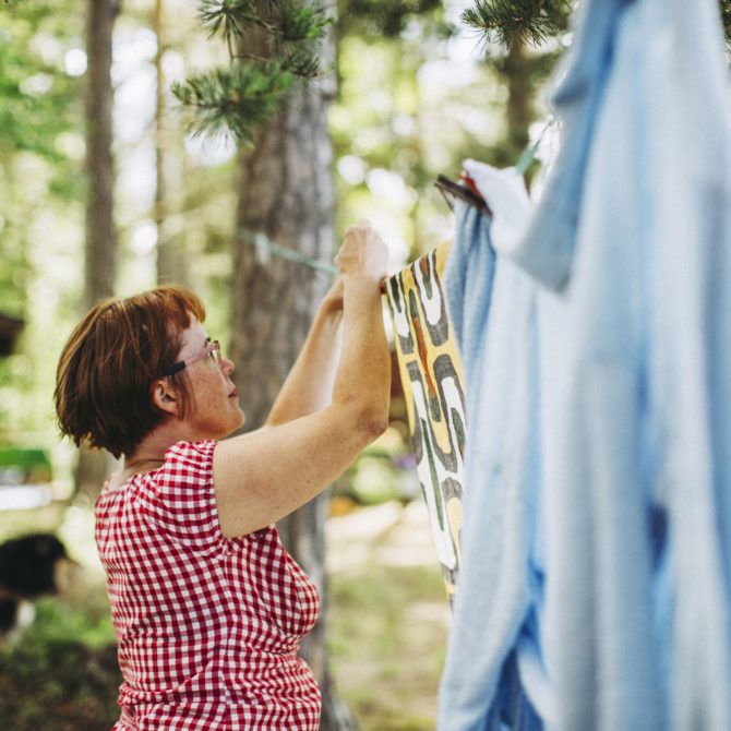 Nainen punavalkoisessa paidassa ripustaa värikästä lakanaa pyykkinaruun, joka roikkuu puussa. Narulle on jo ripustettu sininen lakana ja taustalla näkyy metsää.