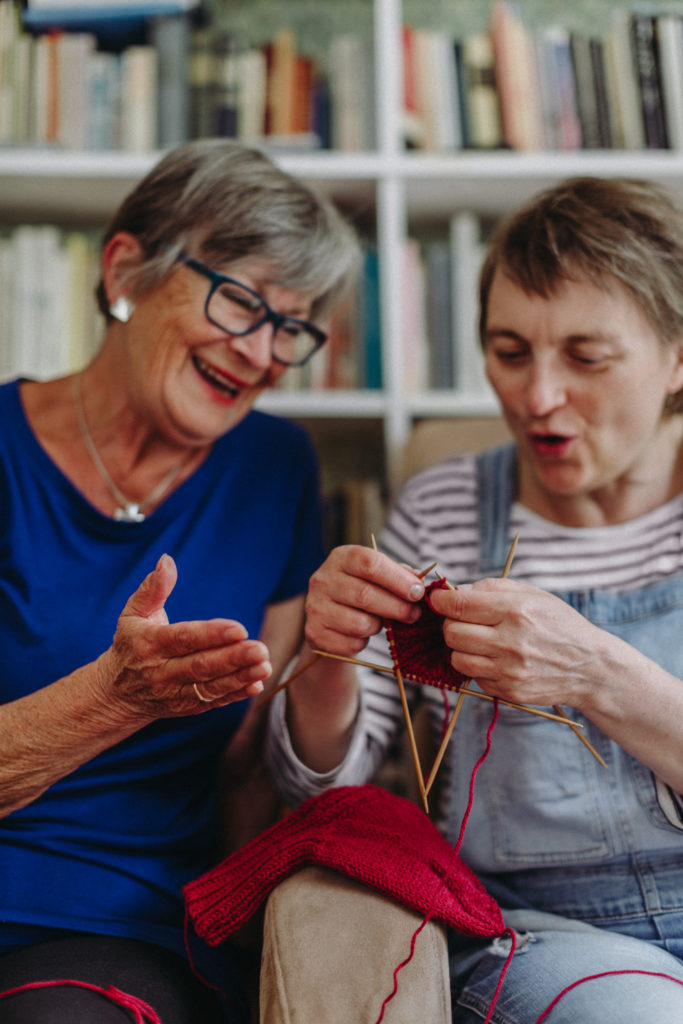 Kaksi iäkästä naista istuvat kirjahyllyn edessä sohvalla. Yksi nainen virkkaa punaisella langalla ja he puhuvat toisilleen hymyillen.