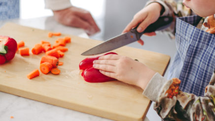Pieni lapsi pitelee paprikaa vaalealla leikkuulaudalla ja leikkaa sitä keittiöveitsellä. Laudalla on pilkottuja porkkanoita ja puolikas paprika ja pöytään nojaa aikuisen käsi. Molemmilla on sininen essu.