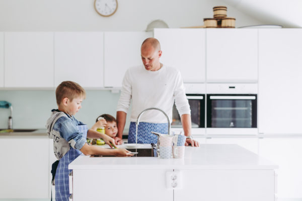 Kaksi lasta ja aikuinen mies seisovat valkoisessa keittiössä vesihanan ympärillä. Yksi lapsi tiskaa lautasta ja toinen nojaa miehen käsivarteen. Kaikilla on siniruudulliset essut päällä.
