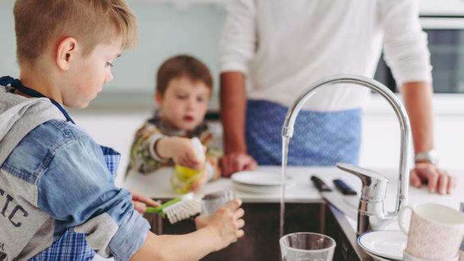 Kaksi lasta ja aikuinen seisovat vesihanan ympärillä siniruudulliset essut päällä. Yksi lapsi tiskaa lasia pesualtaassa ja toinen lapsi suihkuttaa siihen pesuainetta. Aikuinen nojaa keittiöpöytään.