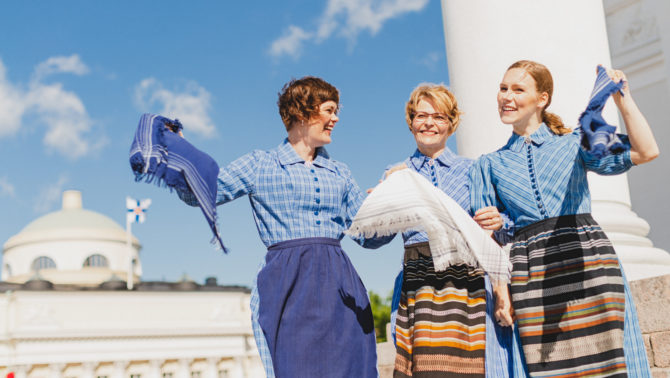 Kolme naista Marttamekoissa seisovat valkoisen pylvään vierellä käsikynkässä ja heiluttelevat pukujensa huiveja tuulessa. Taustalla on sininen taivas ja suuri valkoinen rakennus. Naiset hymyilevät suuresti.