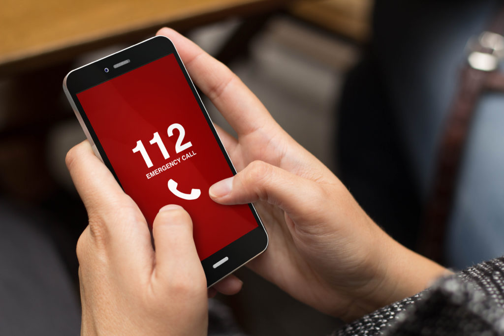 Kädet pitelevät puhelinta, jonka ruutu on punainen ja jossa lukee "112 emergency call".
