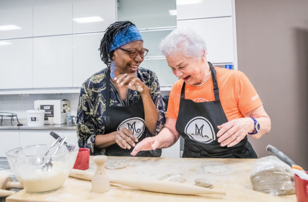 Tumma nainen ja vaalea harmaahiuksinen nainen marttaessuissa leipovat ja nauravat jauhotetun pöydän ääressä.
