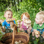 Kolme pientä lasta kyykkivät ruohikossa sienikorin vieressä. Kaikilla on kaksi sientä käsissään ja yksi lapsi nauraa.