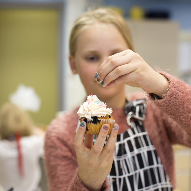 Lapsi mustavalkoisessa essussa pitelee muffinssia ja ripottelee siihen sateenkaariströsseleitä.