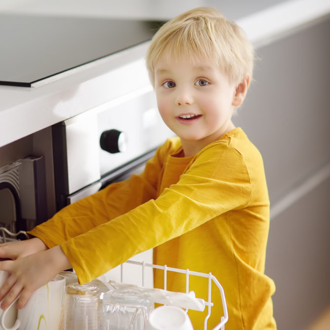 Pieni lapsi keltaisessa paidassa laittaa kahvikuppia tiskikoneeseen.