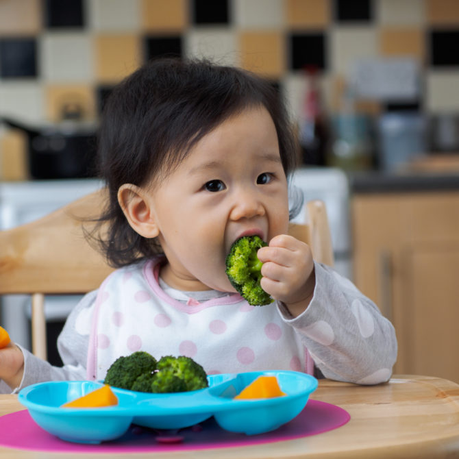 Mustatukkainen vauva istuu keittiössä syöttötuolissa ja syö parsakaalia. Hänen edessä on sininen lautanen, jossa on melonia ja parsakaalia.