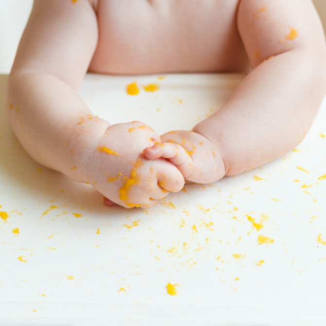 Vauvan kädet syöttötuolin pöydällä. Pöydällä ja käsillä on keltaisia ruoantahroja.