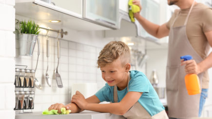 Lapsi ja mies pyyhkivät keittiöpöytää ja kaappeja vihreillä räteillä. Miehellä on toisessa kädessä oranssi suihkepullo ja keittiö on valoisa.