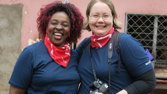 Kaksi naista seisovat hymyillen kuluneen vaaleanpunaisen rakennuksen edessä. Molemmilla on punainen Martat -huivi ja Sininen Martat -teepaita päällä, ja oikeanpuolimmaisella naisella roikkuu kamera kaulan ympärillä.
