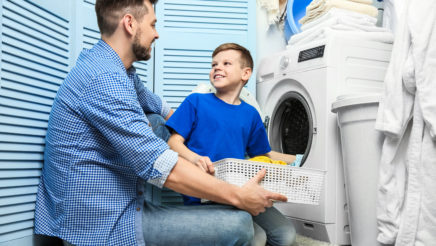 Aikuinen mies ja lapsi istuvat pyykinpesukoneen vieressä ja hymyilevät toisilleen. Miehellä on käsissään pyykkikori ja lapsi laittaa åyykkiä koneeseen.