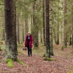 Nainen punaisessa takissa kävelee läpi vihreän metsän pieni kori kädessään. Maa on pudonneiden lehtien peitossa.