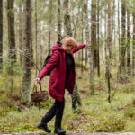 Nainen punaisessa takissa tasapainottelee kaatuneen puun rungon päällä vihreässä metsässä. Hänellä on kädessään pieni ruskea kori ja vapaa käsi on ojennettu sivulle.
