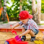 Pieni lapsi punaisessa lippiksessä leikkii hiekkalaatikolla lapiolla ja ämpärillä.