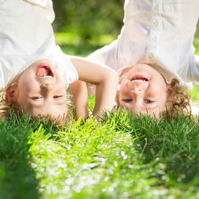 Kaksi pientä vaaleatukkaista lasta valkoisissa paidoissa seisovat päällään nurmella ja nauravat.