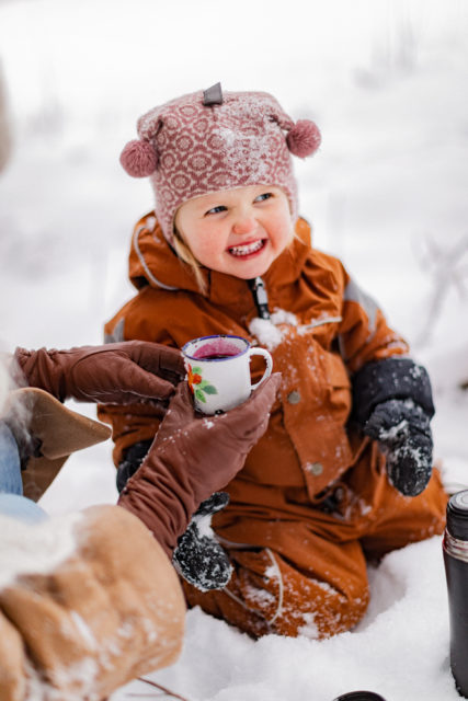 Pieni lapsi oranssissa ulkohaalarissa istuu lumessa ja irvistää. Aikuisen kädet, joissa on nahkahanskat, ojentavat lapselle pientä mukia täynnä mustikkamehua.