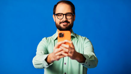 Mies turkoosissa kauluspaidassa seisoo vasten sinistä taustaa. Hänellä on käsissään oranssi puhelin ja pitelee sitä rinnan korkeudella.