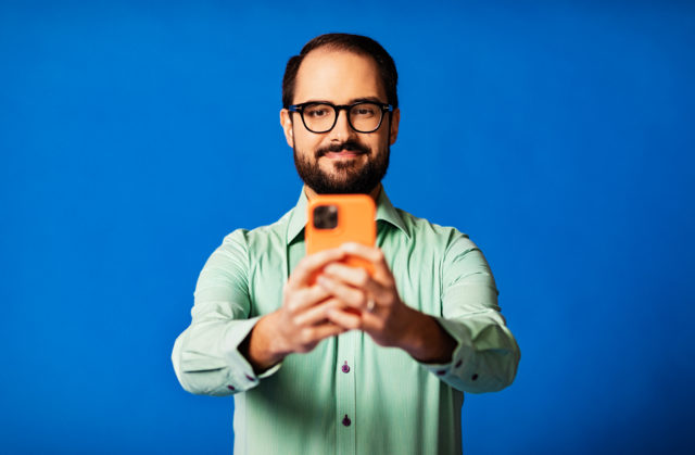 Mies turkoosissa kauluspaidassa seisoo vasten sinistä taustaa. Hänellä on käsissään oranssi puhelin ja pitelee sitä rinnan korkeudella.