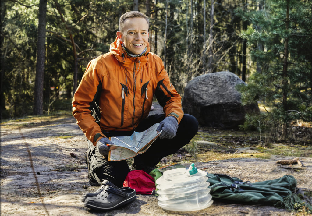 Mies istuu kyykyssä metsässä kallioisella maalla. Hänellä on päällä oranssi takki ja ksäissään kartta, ja hänen edessä on retkeilytavaraa. Takana näkyy puita ja suuri kivi.