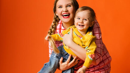 Nainen vaaleanpunaisessa ruudullisessa mekossa nostaa pientä lasta, jolla on keltainen villapaita ja farkut. Molemmat ihmiset nauravat ja tausta on oranssi.