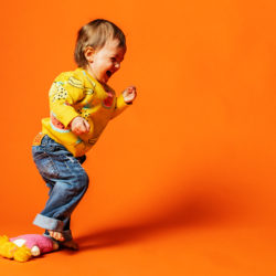 Pieni lapsi keltaisessa villapaidassa ja farkuissa vasten oranssia taustaa. Lapsi nauraa ja nostaa oikeaa jalkaansa, jonka alla on neulottu keltahiuksinen nukke.