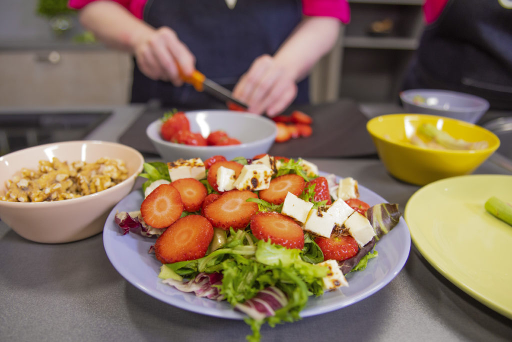 Keittiöpöydällä on lautanen, jossa on mansikka-leipäjuustosalaattia. Lautasen ympärillä on erivärisiä kulhoja ja öeikkuulauta, jonka päällä ihminen pilkkoo mansikoita.