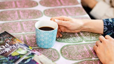 Pöydän päällä on värikäs pöytäliina, kaksi värikästä kirjaa ja kasa paperia. Pöydän ääressä istuu kaksi ihmistä ja molempien edessä on kahvikuppi.