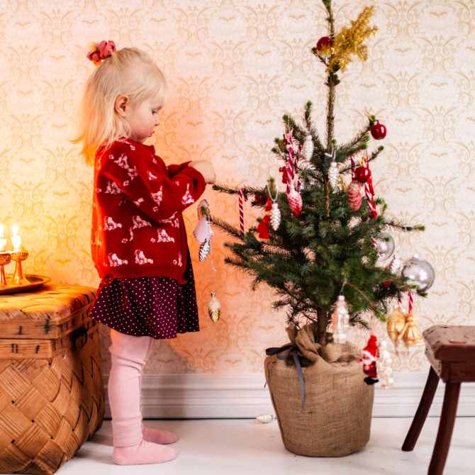 Pieni lapsi punaisessa neuleessa koristelee pientä joulukuusta. Hänen takanaan on olkinen kori, jonka kannen päällä on metallinen viiden kynttilän kynttelikkö, jossa palaa neljä kynttilää. Kuusen takana on jakkara.