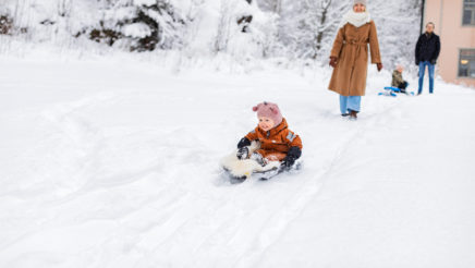 Pieni lapsi oransissa haalarissa istuu pulkassa lumipeitteisessä maassa. Taustalla on kaksi aikuista, toinen lapsi, talon kulma ja luminen maisema.