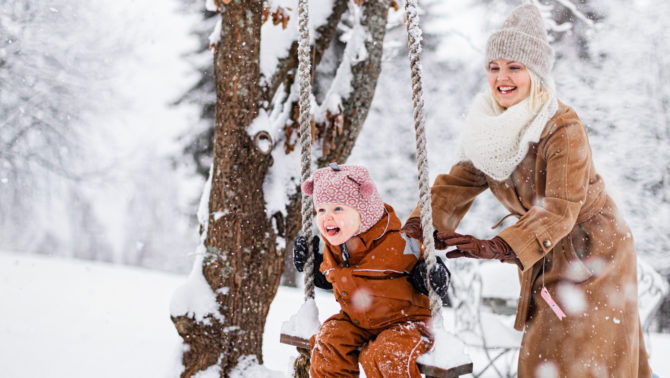 Nainen työntää lasta lumisessa keinussa, joka riippuu puusta. Maa on lumipeitossa ja taustalla näkyy lumisia puita.
