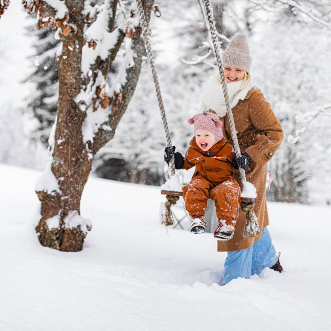Nainen työntää lasta lumisessa keinussa, joka riippuu puusta. Maa on lumipeitossa ja taustalla näkyy lumisia puita.