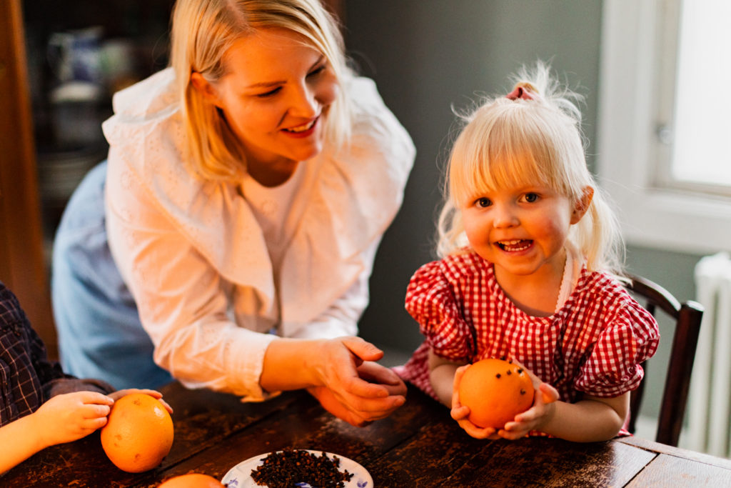Pieni lapsi punaisessa mekossa pitelee neilikka-appelsiinia ja hymyilee. Hän istuupöydän ääressä ja hänen vieressä seisova aikuinen kurkottaa sitä appelsiinia. Toisen pienen lapsen kädet näkyvät pöydän reunalla ja pitelevät omaa appelsiinia.
