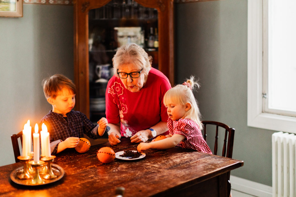 Iäkäs nainen ja kaksi pientää lasta istuvat pöydän ääressä siniharmaassa huoneessa. Pöydällä on palavia kynttilöitä ja nainen auttaa lapsia tekemään neilikka-appelsiineja.