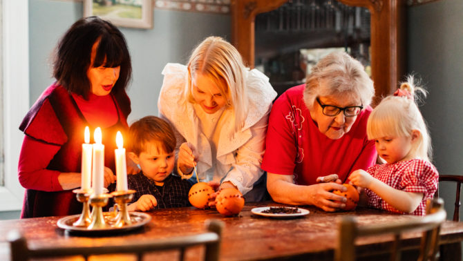 Kolme aikuista naista ja kaksi pientä lasta seisovat pöydän ääressä. Aikuiset auttavat lapsia tekemään neilikka-appelsiineja ja pöydällä on kynttilöitä..