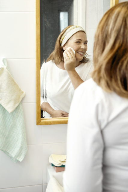 Vaaleahiuksinen nainen seisoo kylpyhuoneessa peilin edessä ja pyyhkii poskeaan pyyhkeellä. Peilin vieressä seinällä roikkuu pyyhkeitä ja nainen hymyilee.