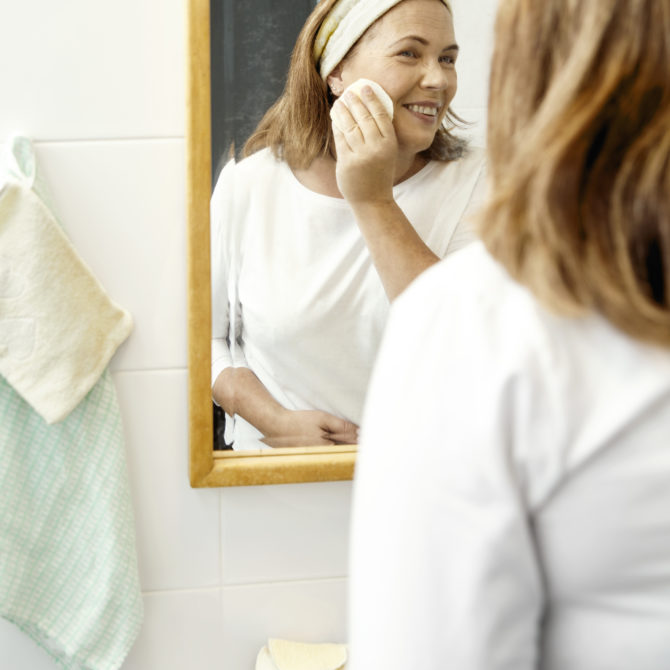 Vaaleahiuksinen nainen seisoo kylpyhuoneessa peilin edessä ja pyyhkii poskeaan pyyhkeellä. Peilin vieressä seinällä roikkuu pyyhkeitä ja nainen hymyilee.