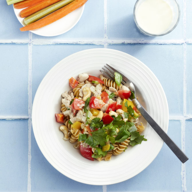 Värikäs pastaruoka sinisellä taustalla valkoisella lautasella, jossa haarukka reunalla. Vieressä maitolasi ja pienellä lautasella tuorekurkkua ja porkkanaa suikaleina