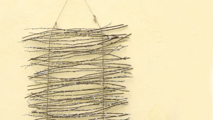 Pajun varvuista koottu verhomainen koriste roikkuu vaaleankeltaisella seinällä. Koristeen alareunassa on neljä pikkunarsissia sipuleineen.