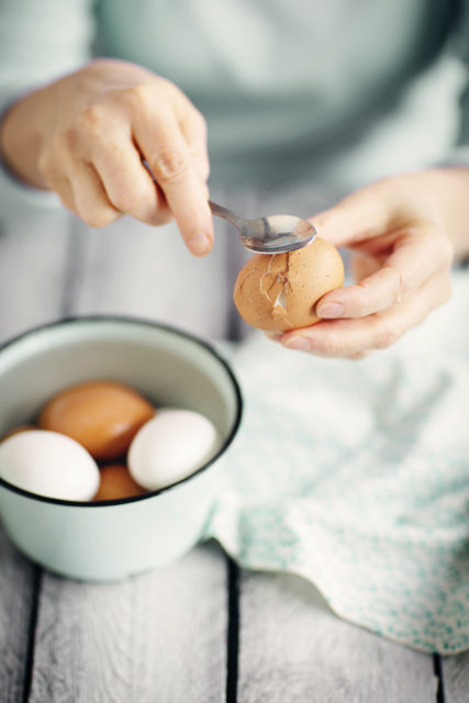 Käsi käyttää teelusikkaa rikkomaan kananmunaa. Taustalla on kulhollinen kananmunia.
