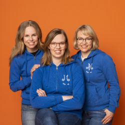 Kolme vaaleahiuksista naista Marttamekoissa poseeraavat vasten oranssia taustaa ja nauravat. Keskimmäinen istuu ja kaksi muuta seisovat hänen ympärillä.