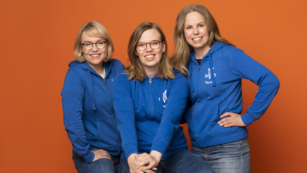 Kolme vaaleahiuksista naista sinisissä Marttaliitto huppareissa istuvat ja hymyilevät vasten oranssia taustaa.