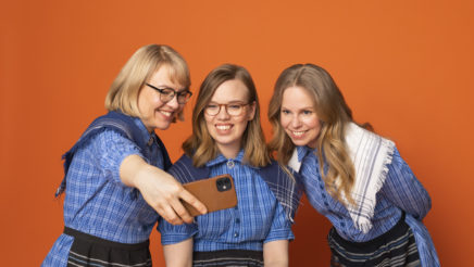 Kolme vaaleahiuksista naista Marttamekoissa ottavat selfietä yhdessä vasten oranssia taustaa.