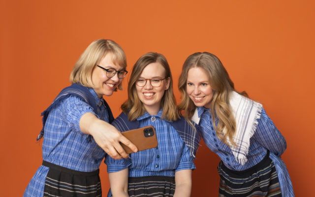 Kolme vaaleahiuksista naista Marttamekoissa ottavat selfietä yhdessä vasten oranssia taustaa.