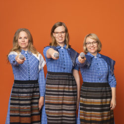 Kolme vaaleahiuksista naista Marttamekoissa seisovat rivissä. Heillä on mikrofonit käsissään ja osoittavat niitä eteenpäin.