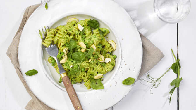 Keväisen vihreä pastaruoka-annos valkoisella syvällä lautasella. Koristeena basilikan lehtiä ja herneen versoja.. Lautasen reunalla puupäinen haarukka. Lautanen pellavakankaan päällä pöydällä.