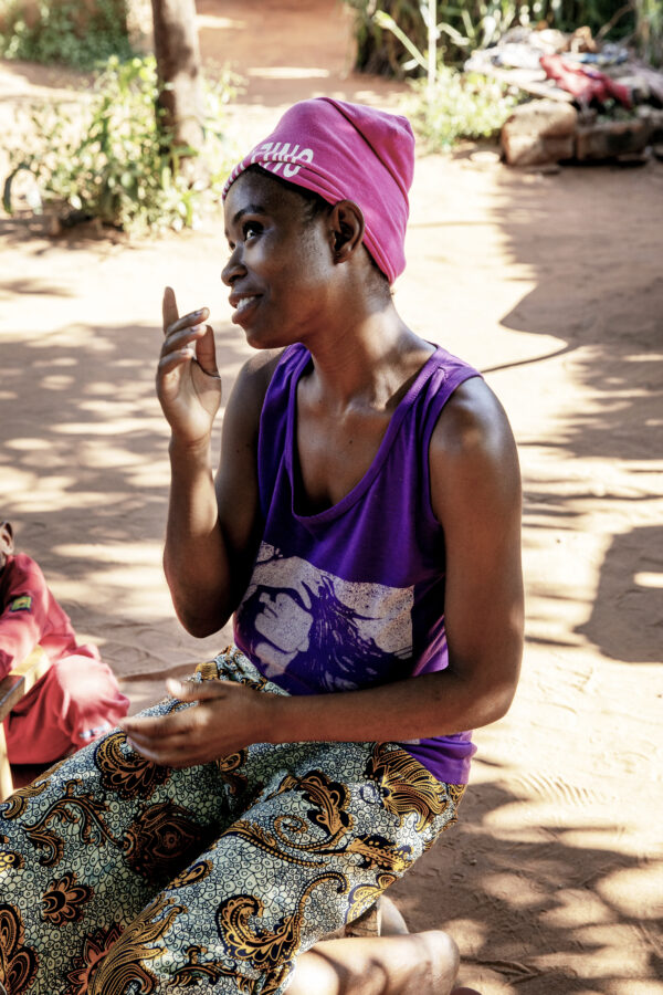 Afrikkalaistaustainen nuori nainen istuu polvillaan maassa, hänellä on voiletti paita, kirjava lannevaate ja pinkki päähine. Hän viittoo ja katsoo ohi kameran kuvassa vasemmalle.